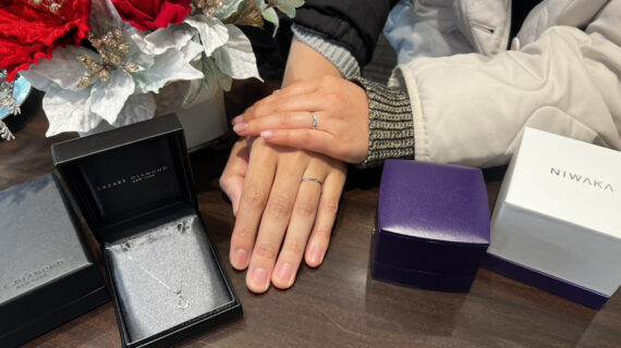 箱に入ったラザールダイヤモンドの婚約ネックレスとNIWAKA（ニワカ）の結婚指輪を着けたカップルの手元